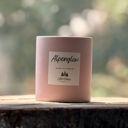 Pink candle labeled 'Alpenglow' by 'Little Pines' on a wooden surface with a blurred natural background.